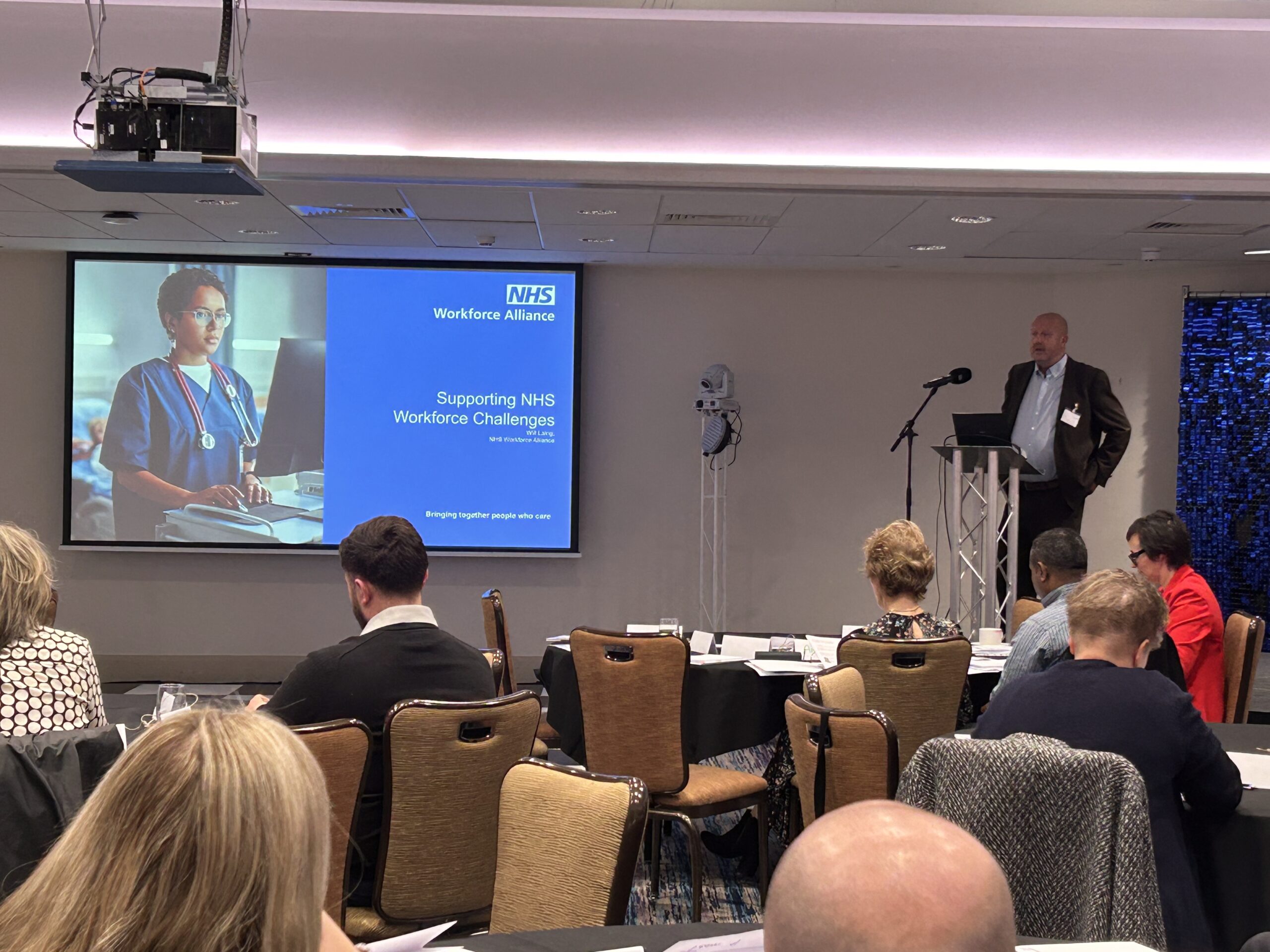 Workforce Alliance Operations Director, Will Laing, stands behind a podium on stage, speaking to an audience at the NHS Workforce Alliance Joint Cluster Workforce Workshop in Sheffield