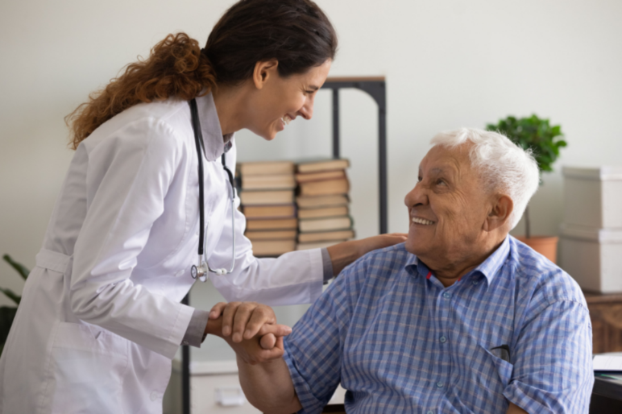 A female health worker in a white coat holds the hand of a seated, elderly male patient who is smiling