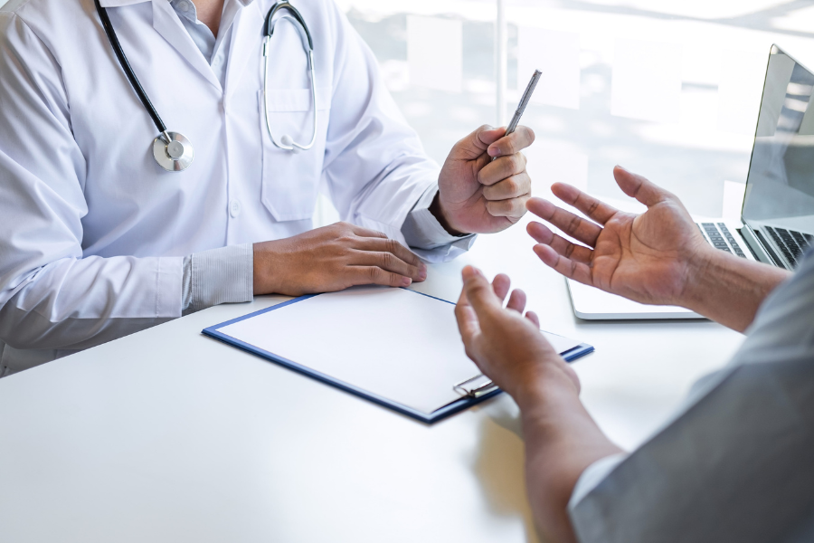 A close-up of two people having a discussion over a table. On the table is a clipboard and one of the people is dressed in a white medical coat and is holding a pen.
